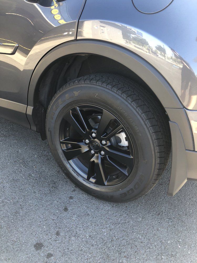 A black car with black wheels and tires is parked on the side of the road.