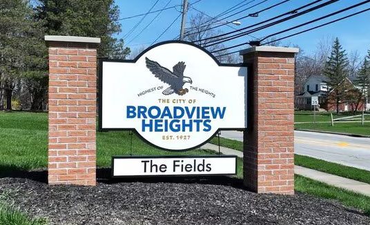 A sign for The Fields in Broadview Heights, featuring a blue and white eagle logo on brick pillars beside a road.
