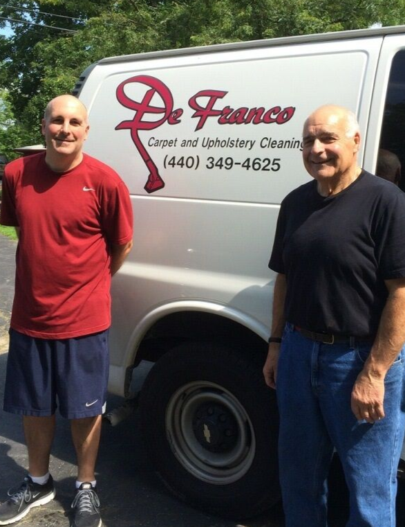 Two men stand outdoors in front of a white work van labeled 