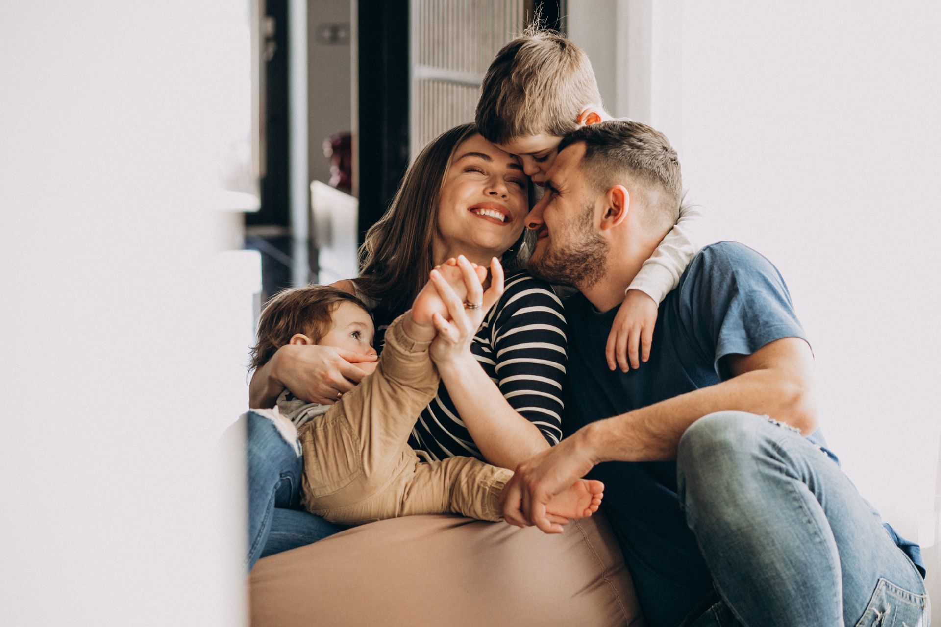 Family of four smiling, sitting together. Woman holds toddler while man embraces her, other child on the man's shoulders.