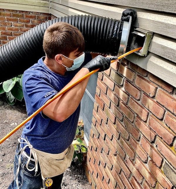Person cleaning a dryer vent outside a brick building, wearing a mask and gloves.