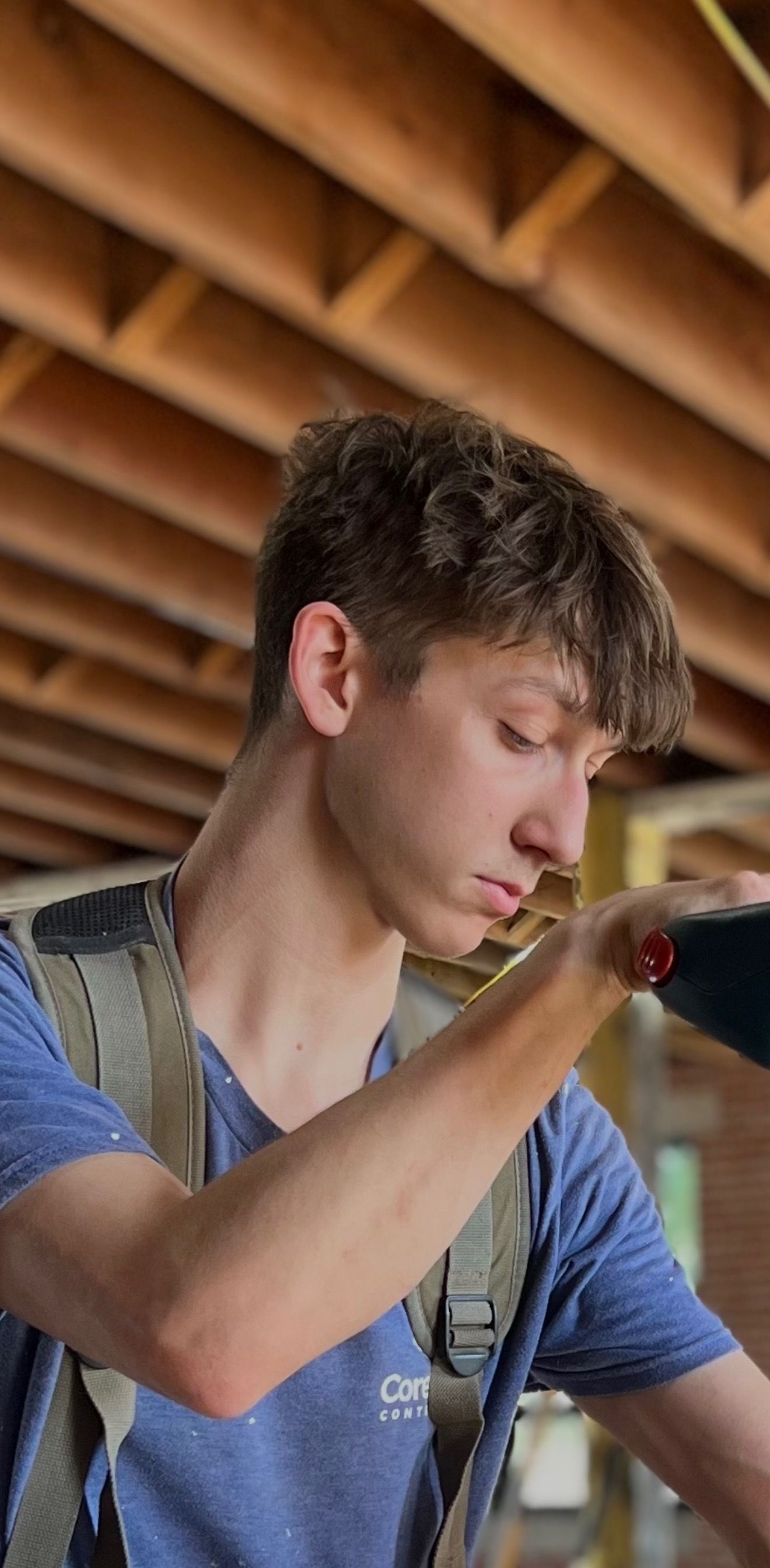 A person with curly hair wearing a blue shirt and work vest, holding a tool, indoors, looking down.