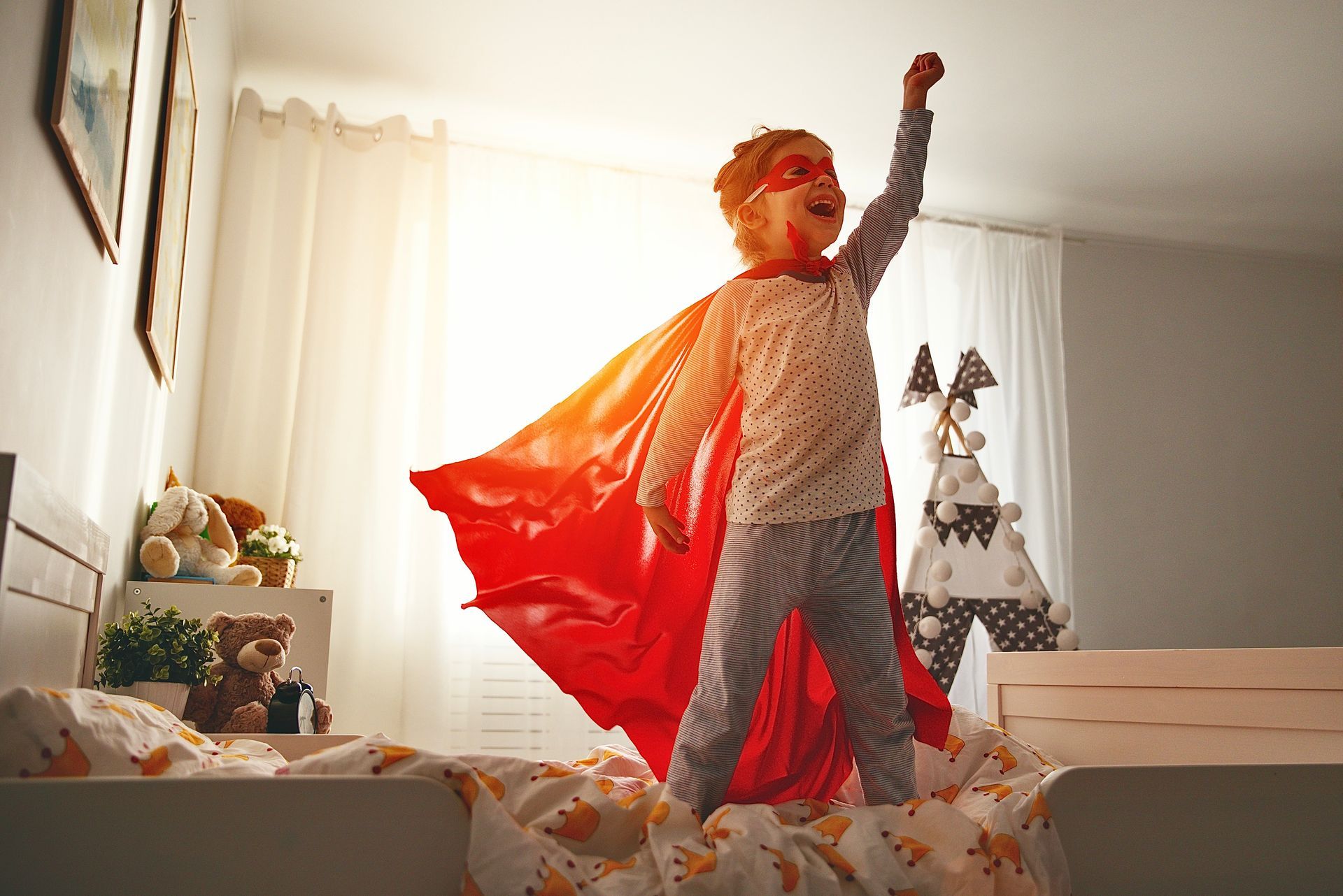 Child in superhero costume, red cape, mask, arm raised victoriously on bed in a bedroom.