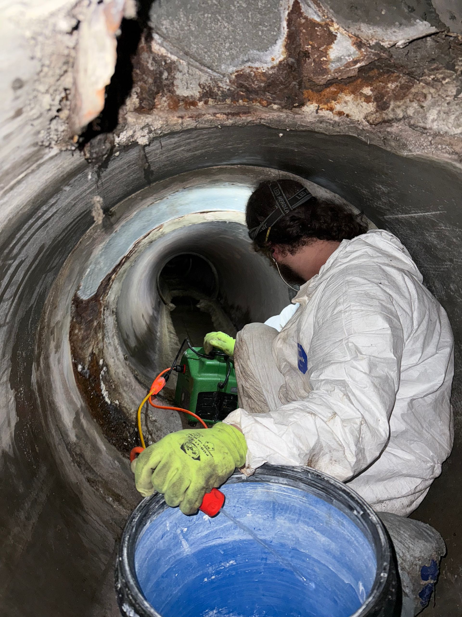 Person in protective suit working inside a concrete pipe with a green machine and bucket.