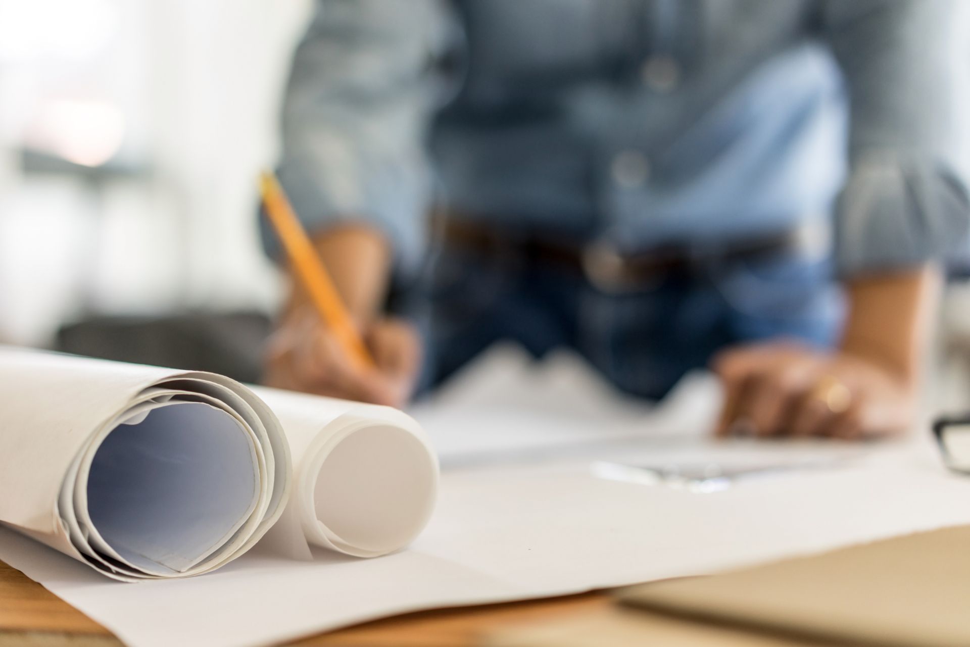 Rolled blueprints in the foreground, blurred person writing on paper with a pencil.