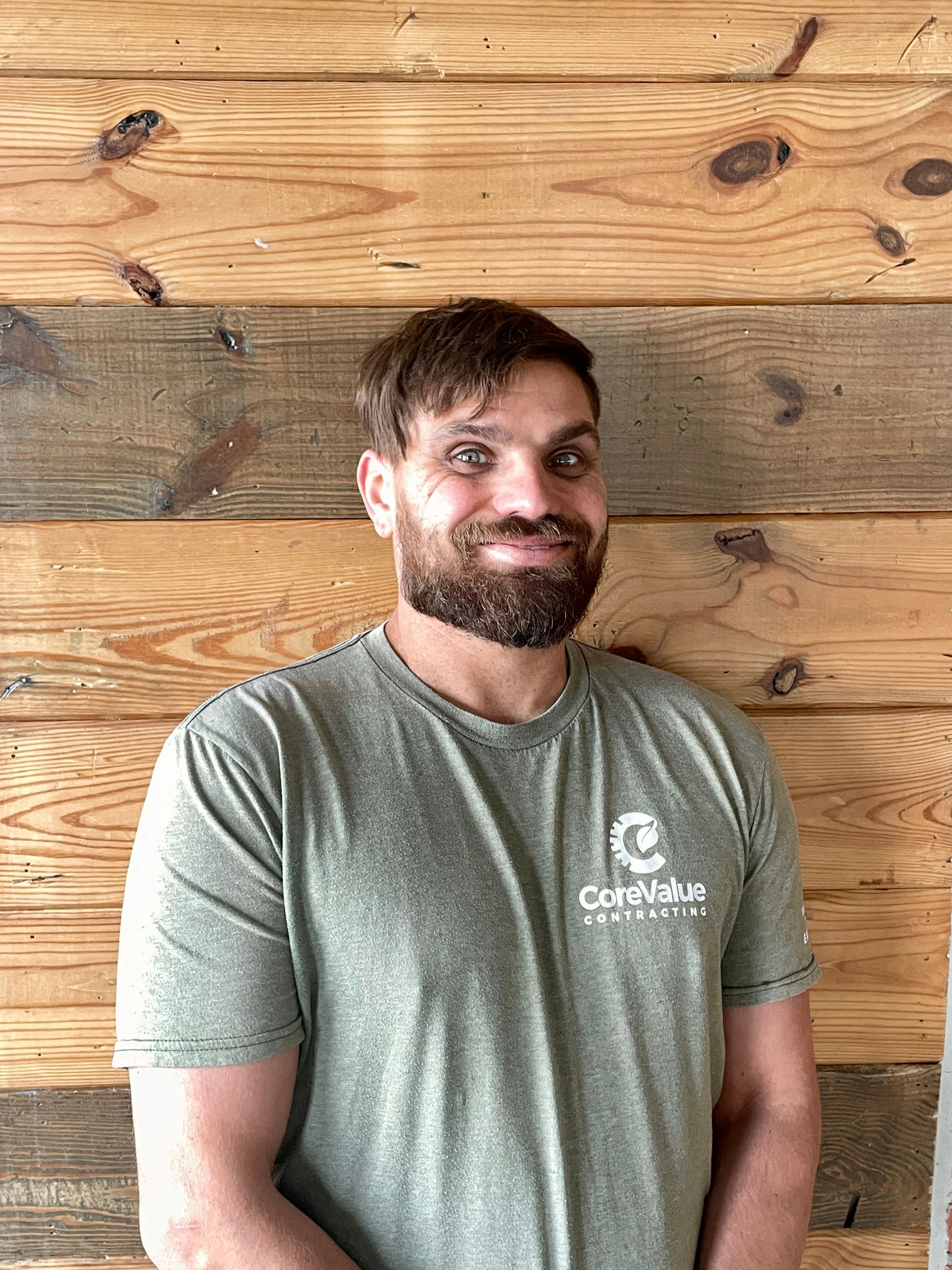 Man with beard, wearing a green t-shirt, smiles in front of a wood-paneled wall.