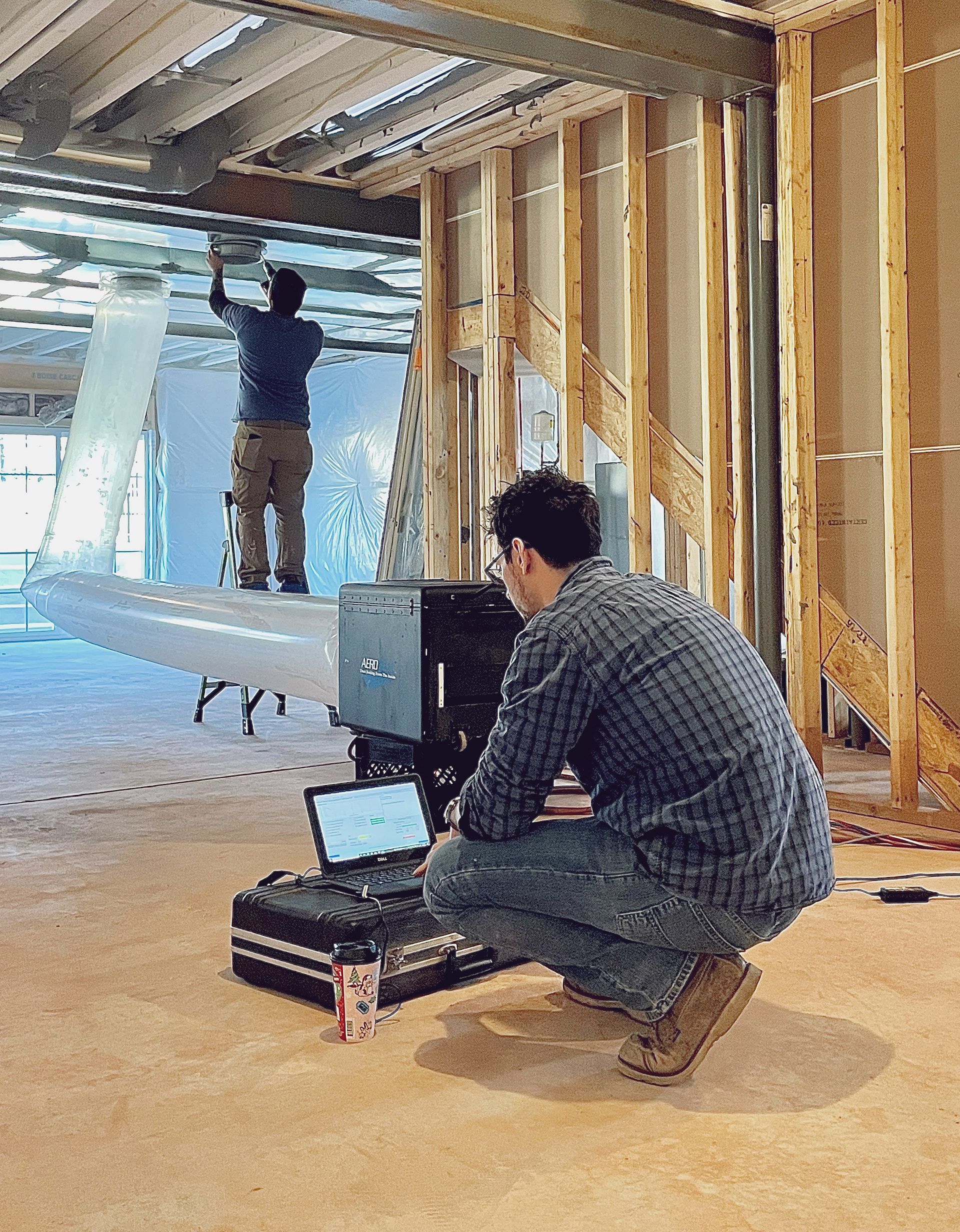 Two construction workers in a building under construction. One on a scaffold, another crouched at a computer.