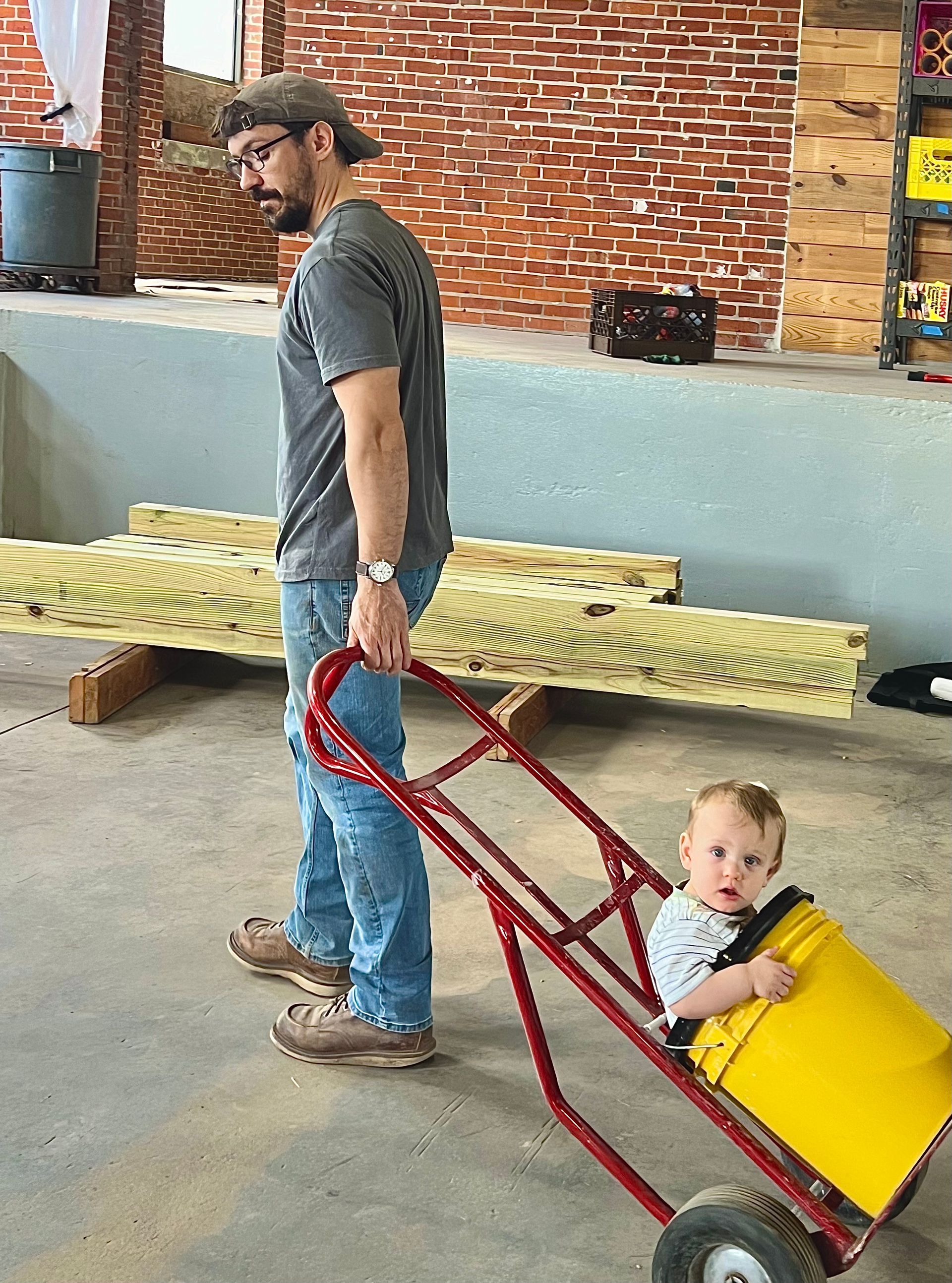 Man pulling a red hand truck with a yellow bucket holding a baby. Man in jeans and a gray shirt.