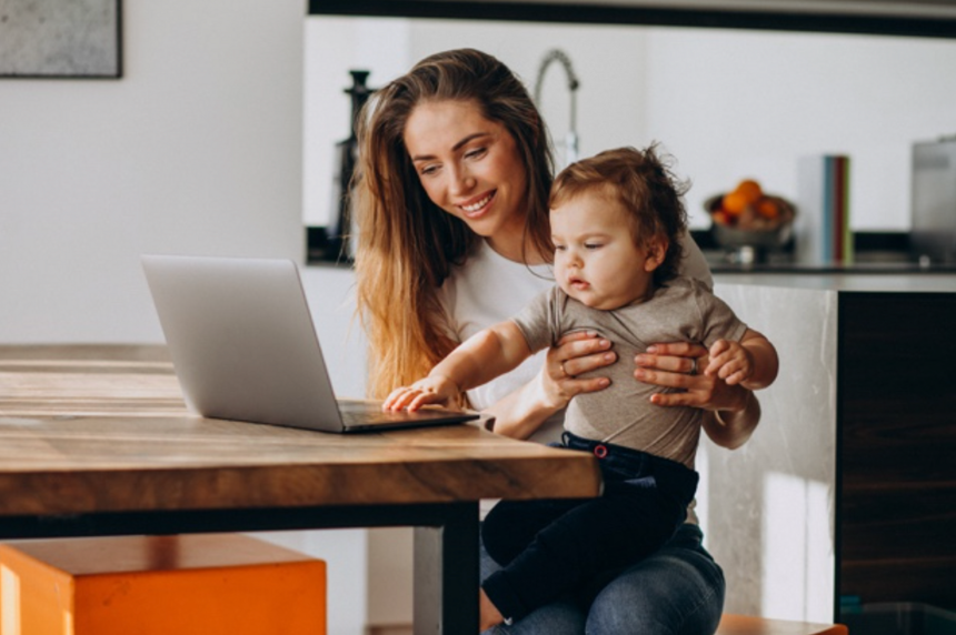 Woman holding baby, using laptop at kitchen table. Baby points at the screen. Bright interior.