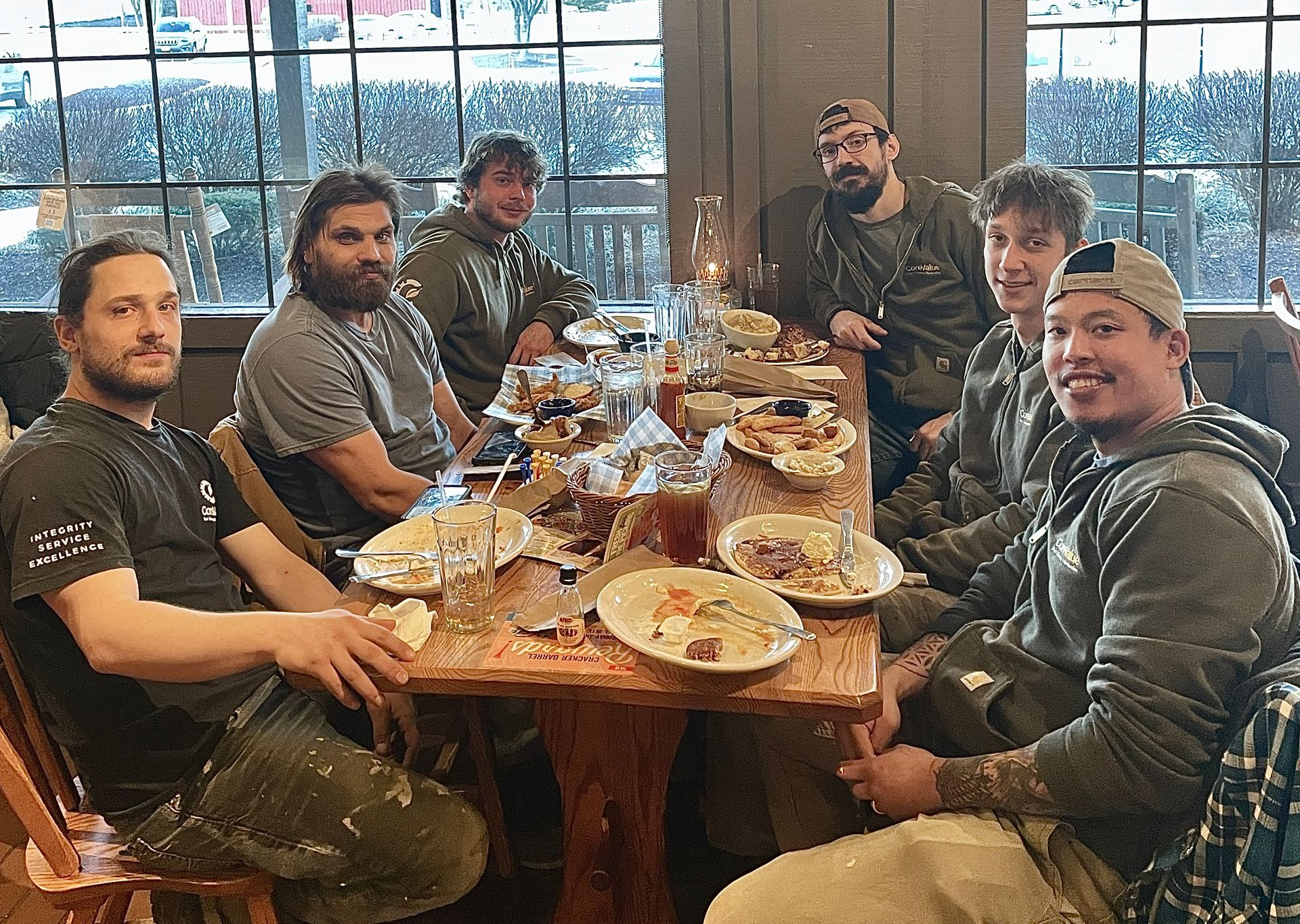 Group of people at a wooden table with food and drinks. Some are smiling, in a restaurant setting.