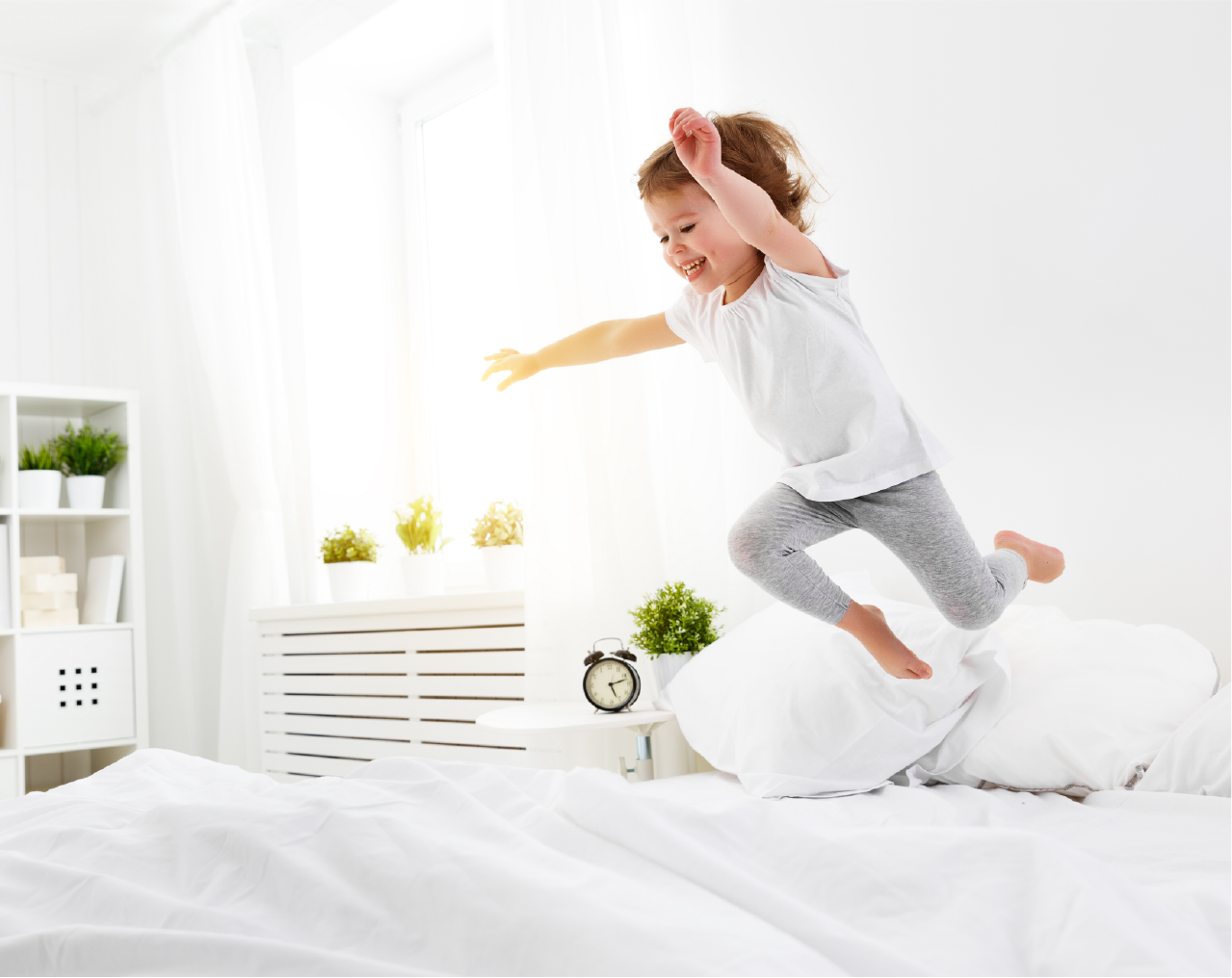 Girl jumps joyfully on a white bed in a bright bedroom, reaching out, smiling.