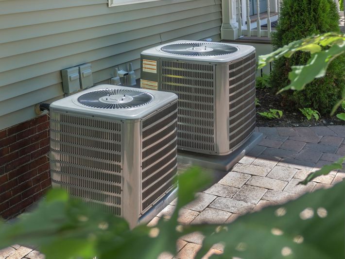 Two air conditioning units on a brick patio next to a light-colored house, surrounded by greenery.