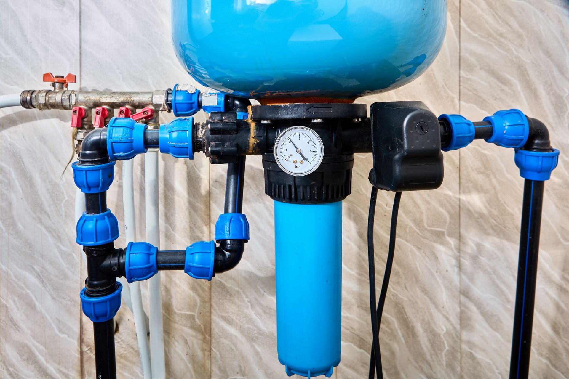 Blue well water filtration system with pressure gauge and PVC fittings against tiled background.
