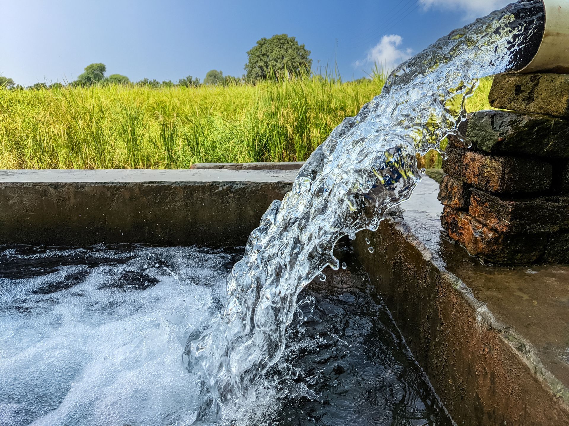 Water flowing from pipe into tank with green fields in background.