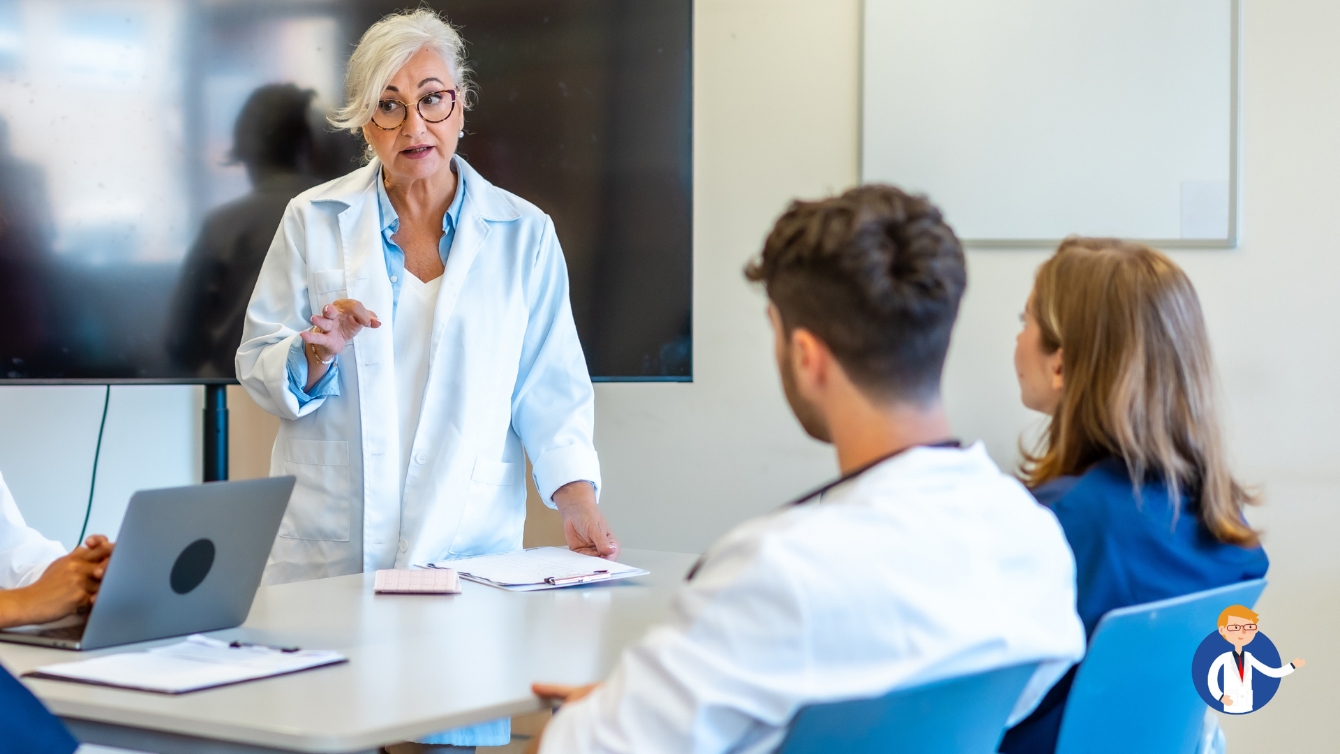 A female doctor in lab coat presents to a seated group. They are in a conference room.