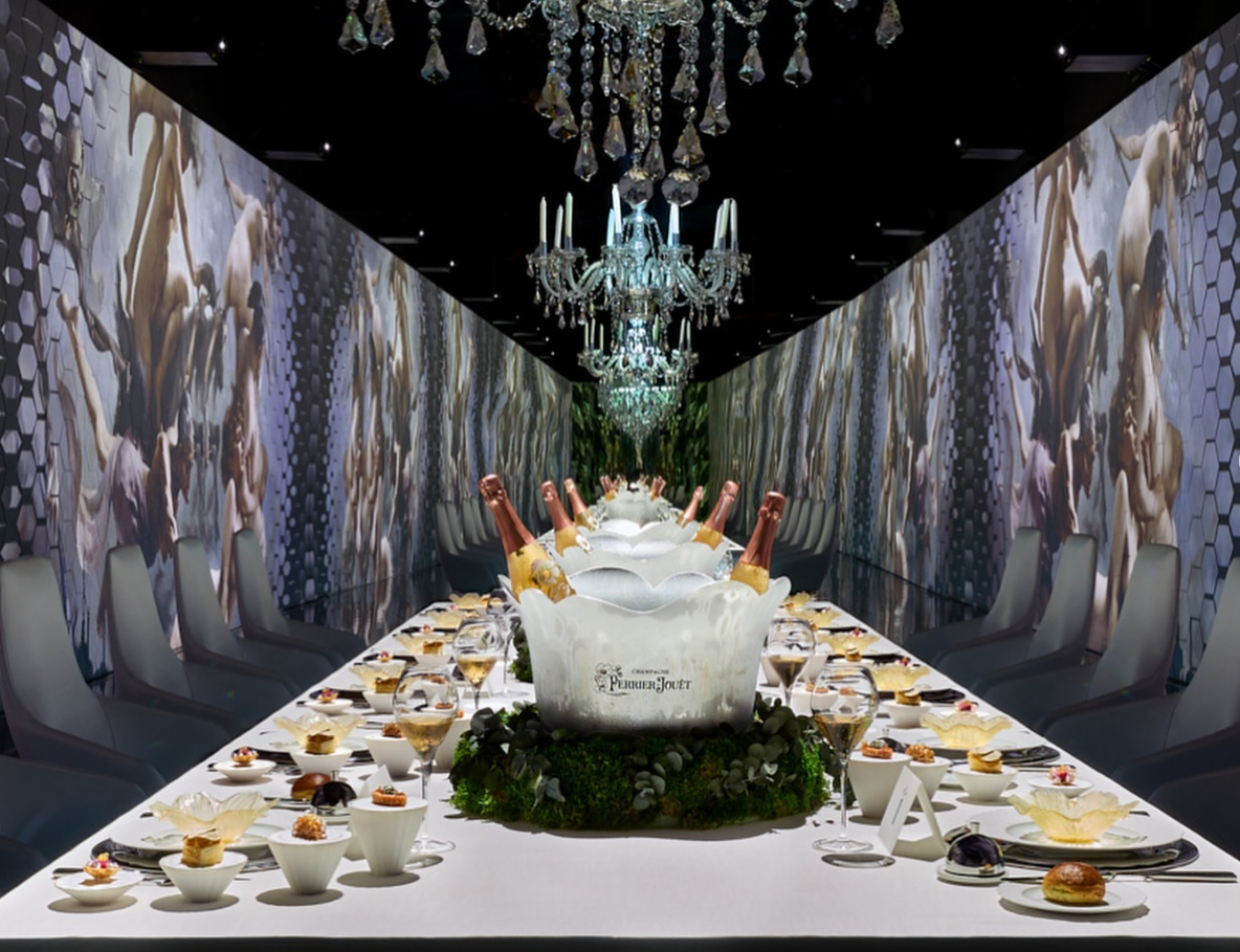 Long dining table set for a formal event, with champagne and food under crystal chandeliers. Walls have abstract art.