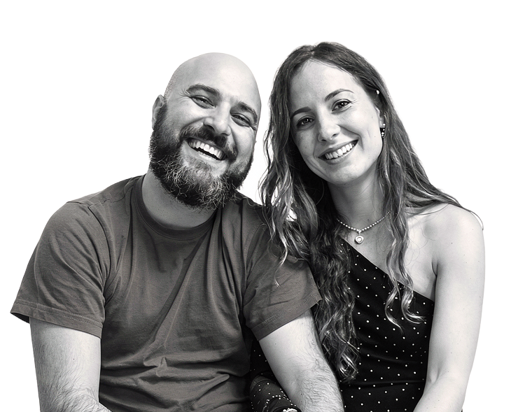 Smiling couple sits close together; man wears a t-shirt and jeans, woman in a one-shoulder black top and lace leggings.