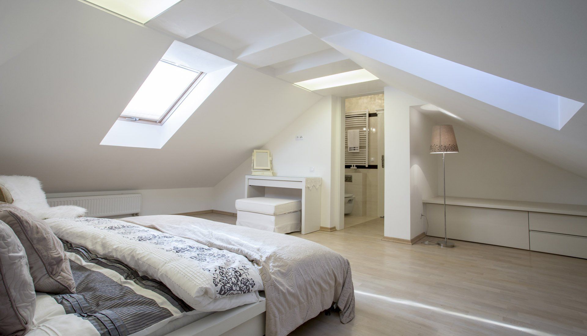 A bright, minimalist attic bedroom featuring a sloping ceiling, a skylight, a queen bed, and a small vanity area.