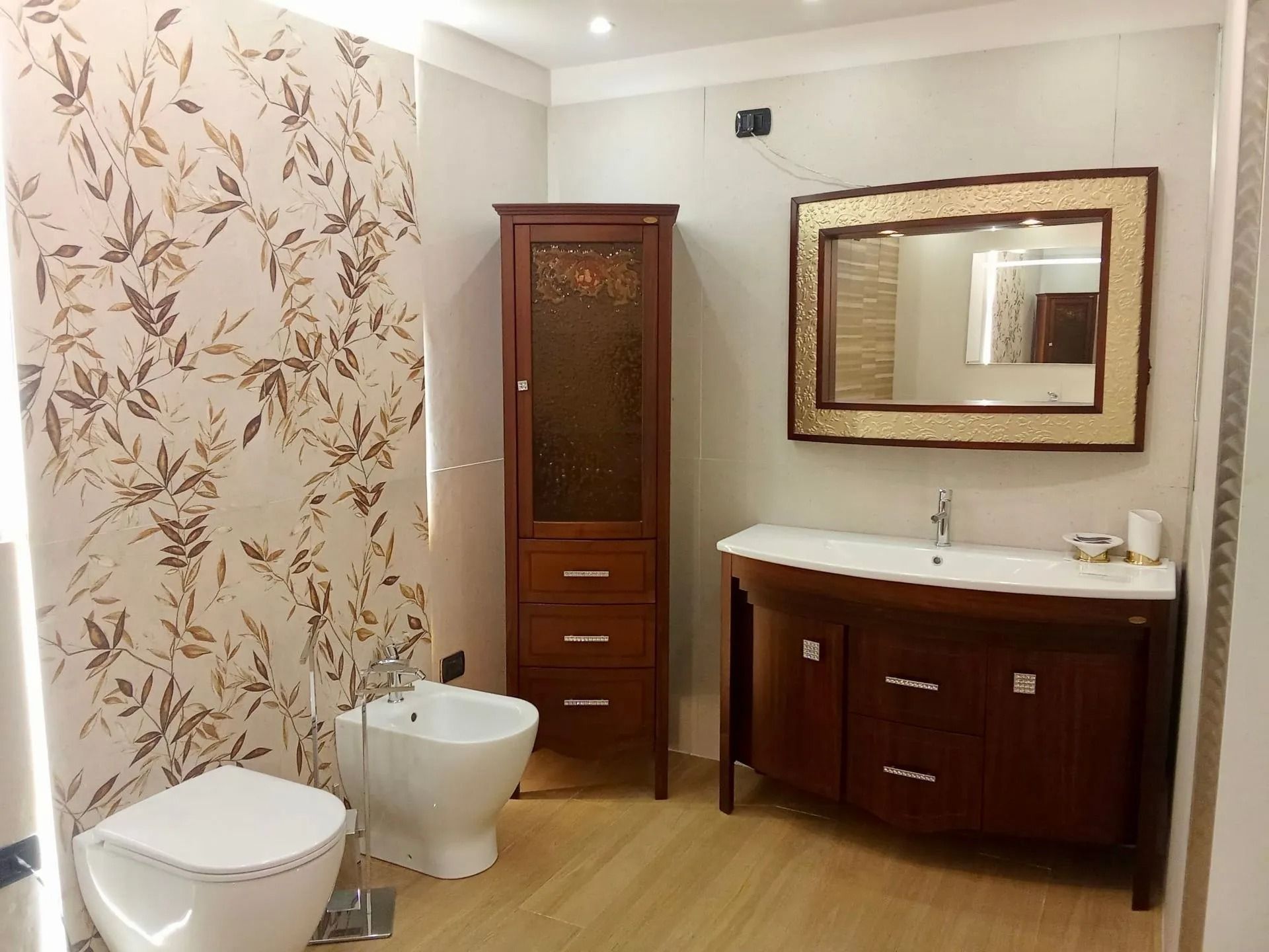Bagno moderno con WC, bidet, lavabo e carta da parati decorativa a motivi floreali.