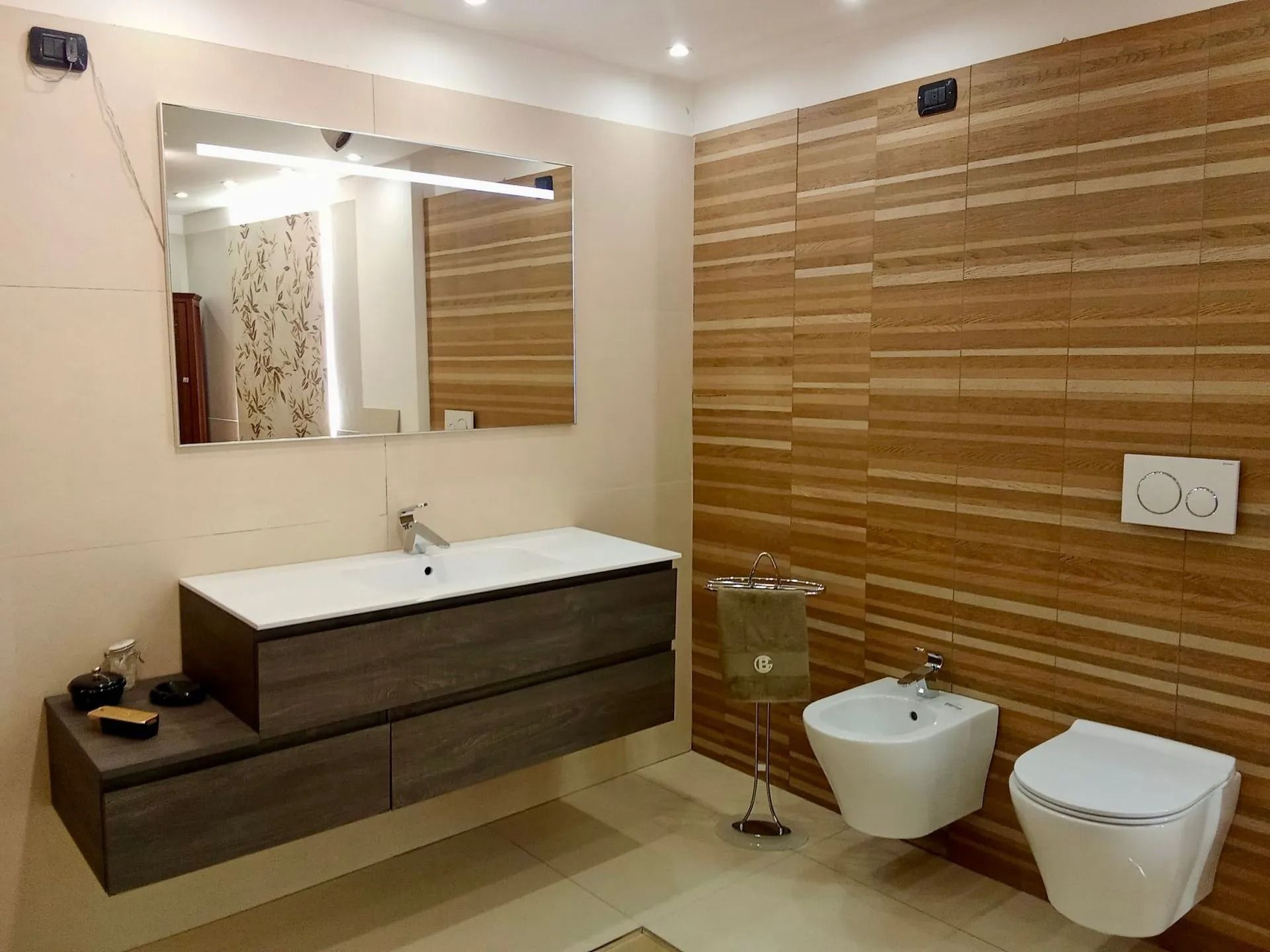 Bagno moderno con rivestimento a parete in piastrelle effetto legno, mobile lavabo sospeso, specchio, box doccia in vetro e due WC sospesi.