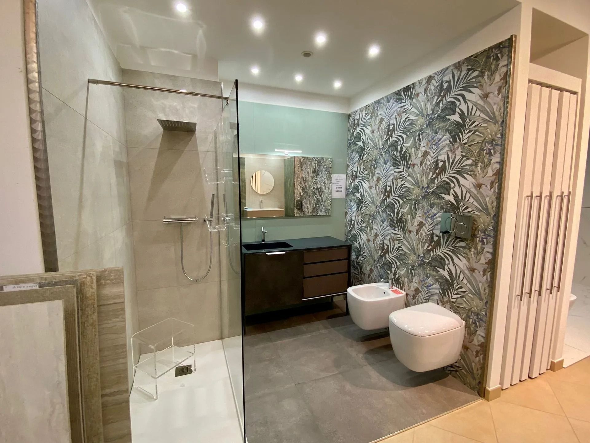 Bagno moderno con doccia in vetro, mobile lavabo scuro, sanitari bianchi e parete decorata con motivi floreali.