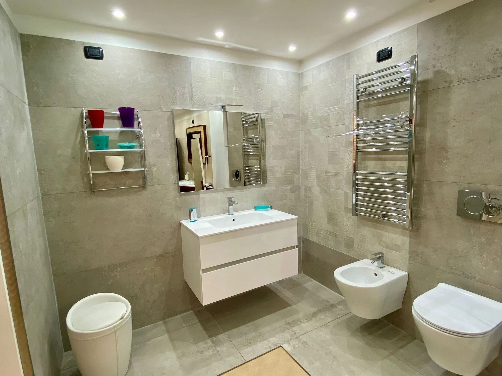 Bagno moderno con piastrelle beige, lavabo a parete, WC, bidet e scaldasalviette cromati.
