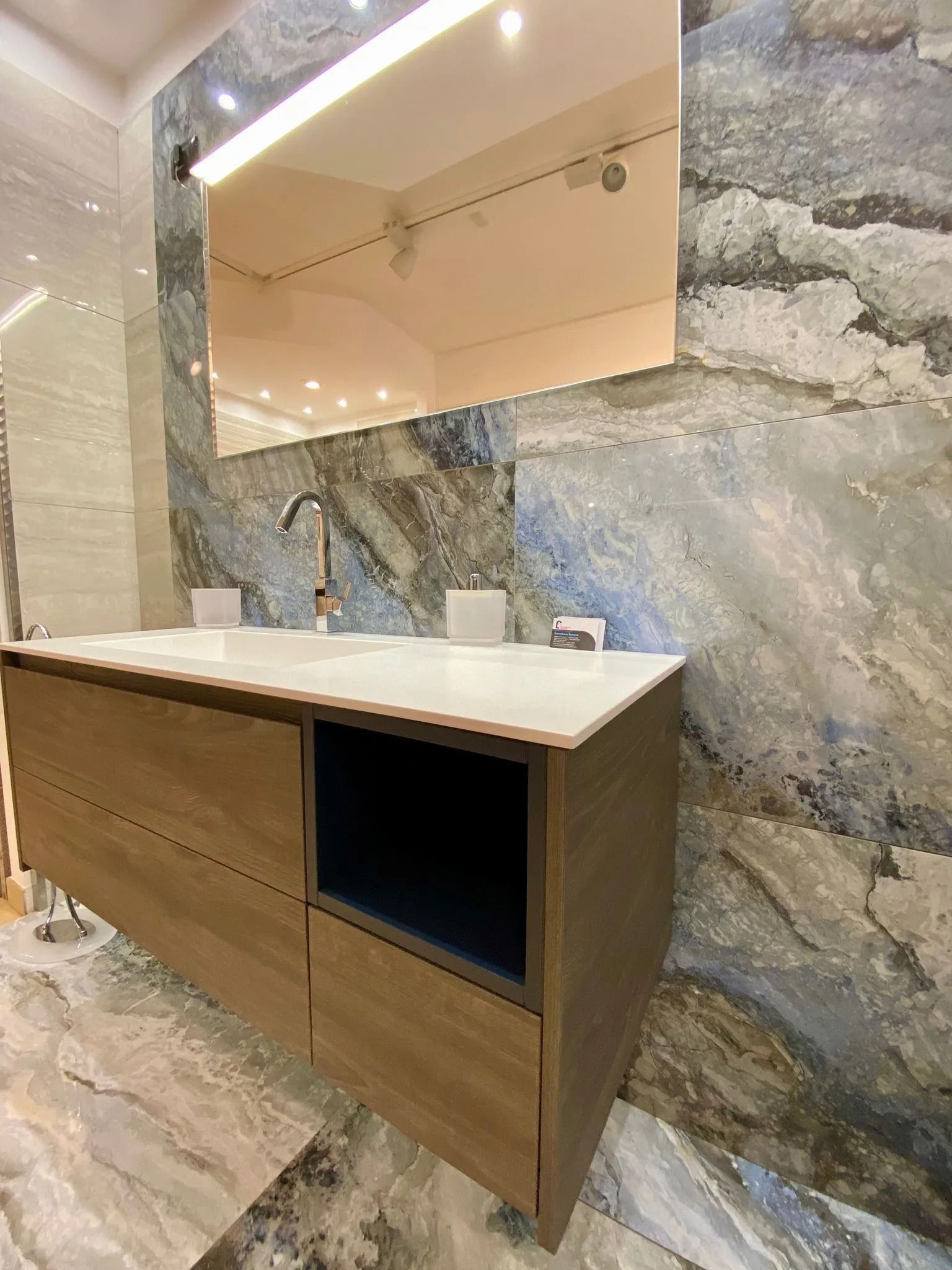 Mobile bagno moderno con mobiletto in legno, piano d'appoggio bianco e grande specchio su una parete in marmo.