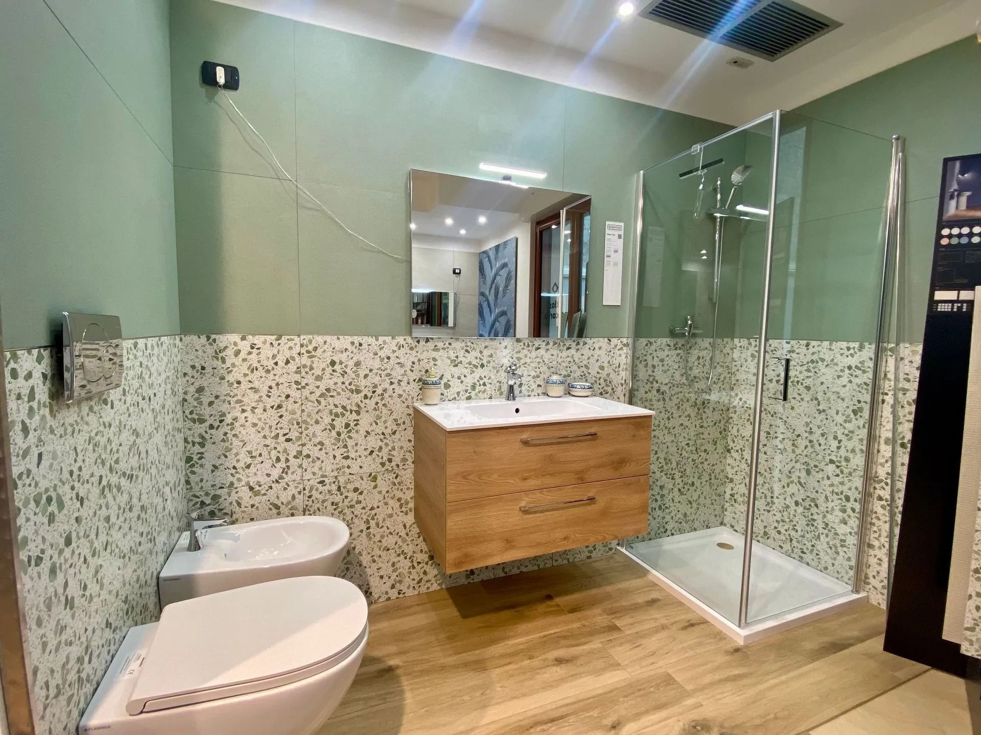 Bagno moderno con doppio lavabo, WC, bidet e doccia con box in vetro, pareti verdi e mobiletto in legno.