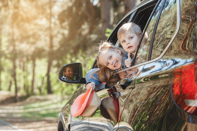 Two children leaning out a car window on a sunny tree-lined road, smiling at the camera