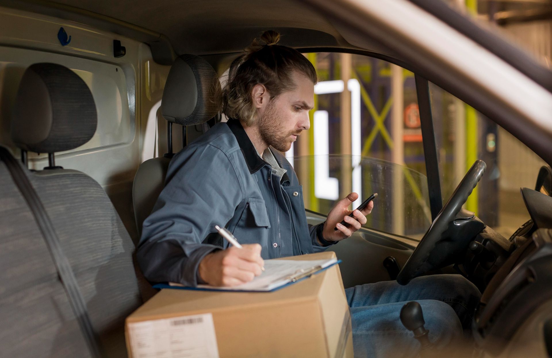 Delivery worker in a van writing on a clipboard while checking a smartphone