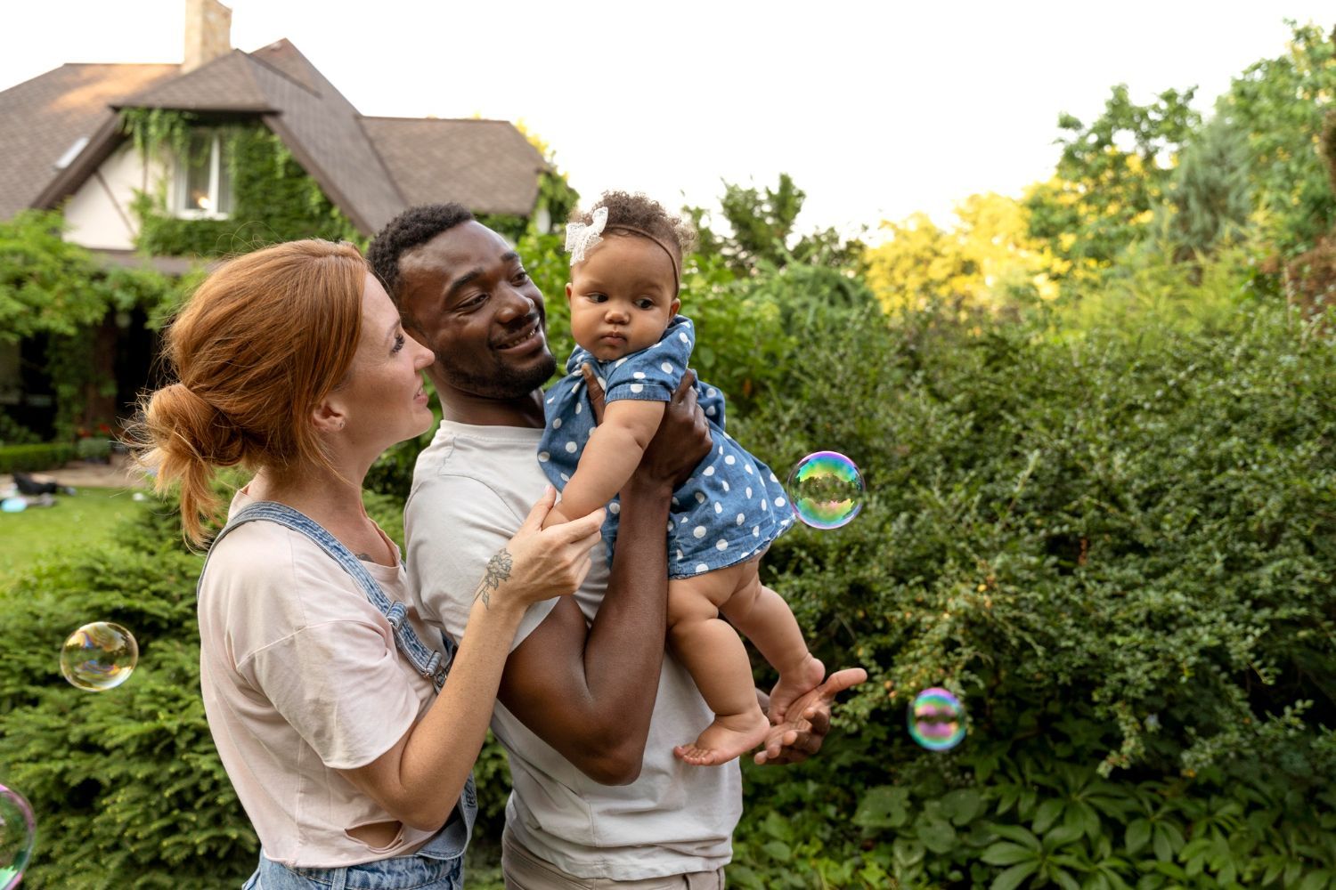 Two adults holding a child outdoors in a garden, with soap bubbles and a house in the background