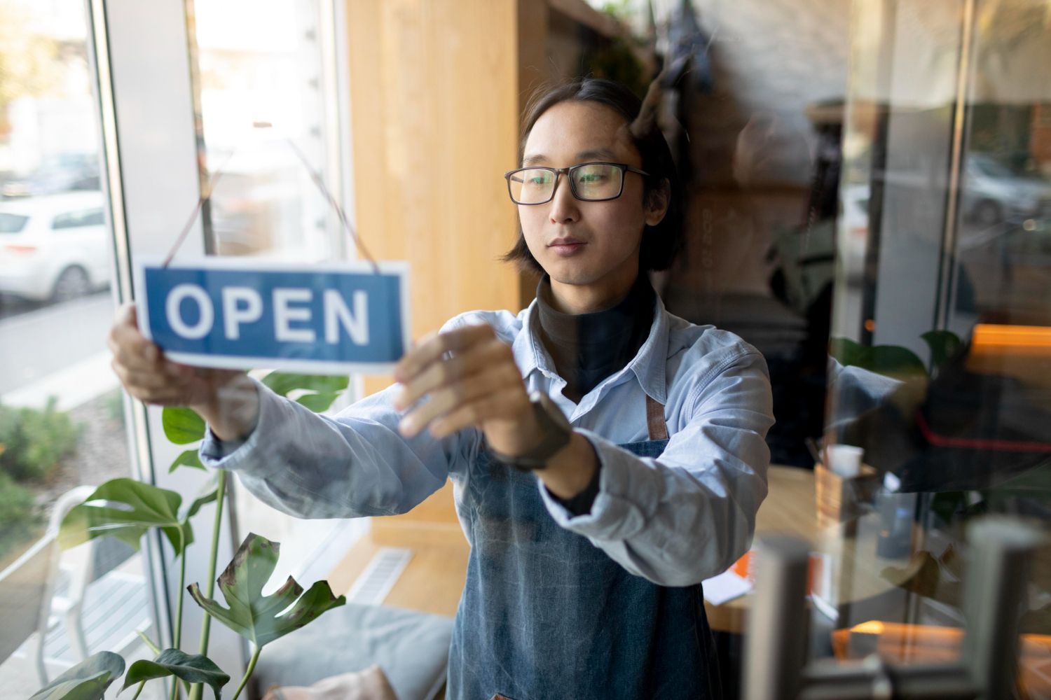 Person hanging an OPEN sign on a glass storefront, wearing glasses and an apron.
