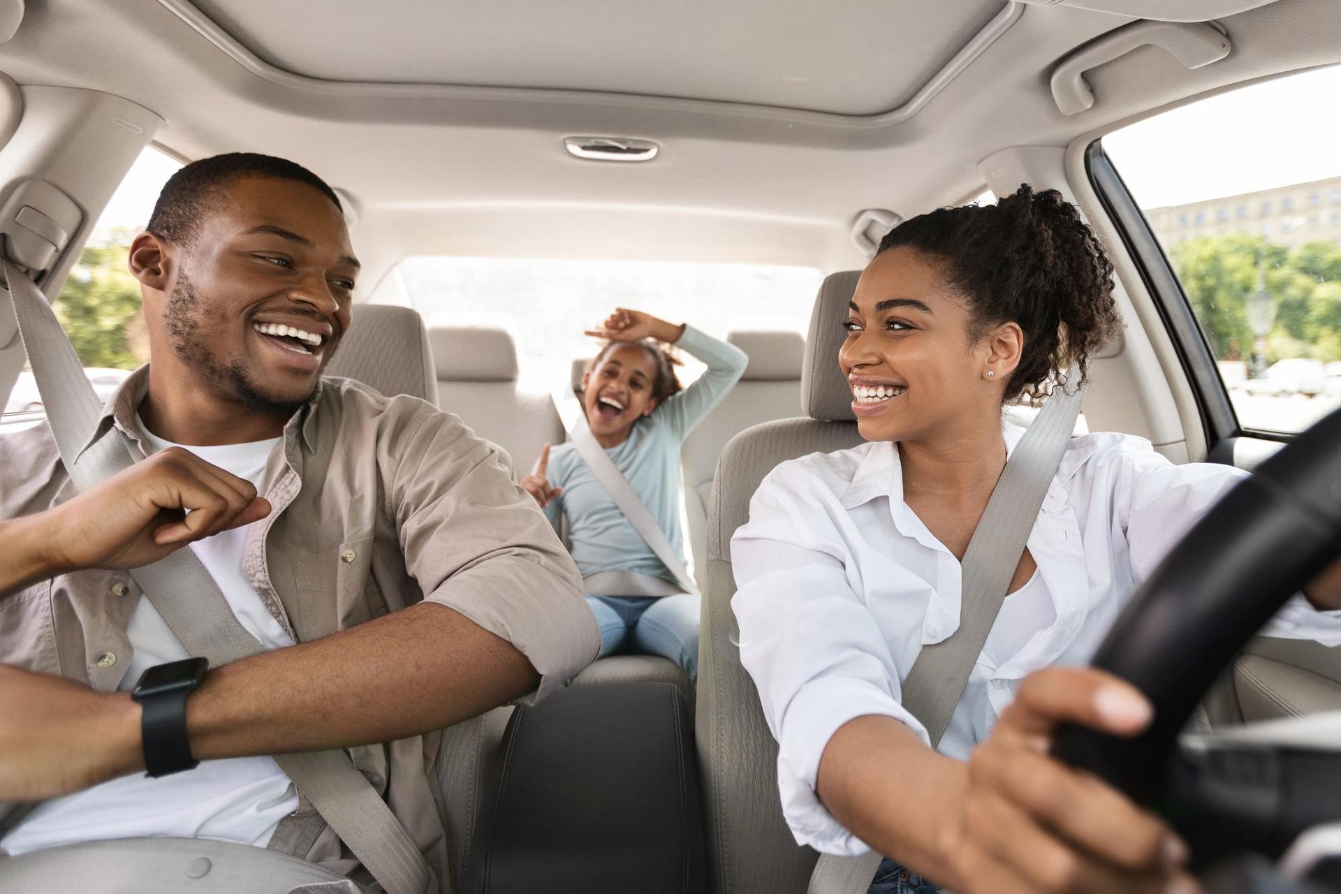 Three friends laughing in a car, with the driver smiling at the front and a child cheering in the back seat.
