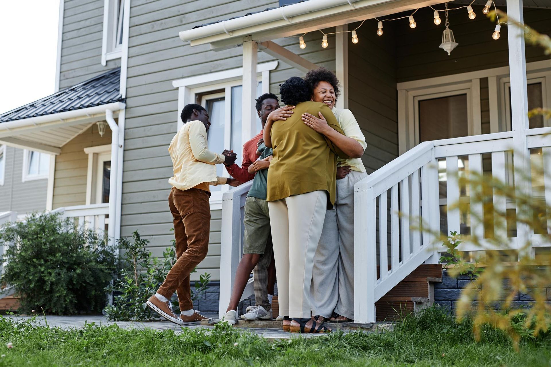 Two people hugging on a front porch while others watch, with a house and string lights in the background