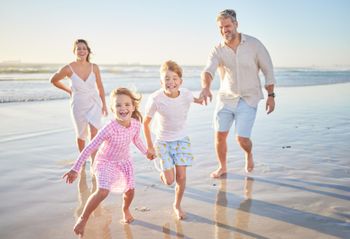 Family running barefoot along a sunny beach at low tide, smiling and splashing water