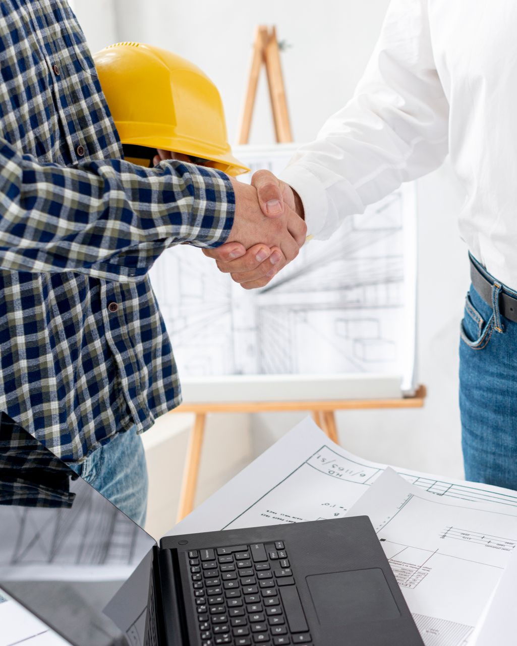 Two workers shaking hands over blueprints with a yellow hard hat and laptop nearby