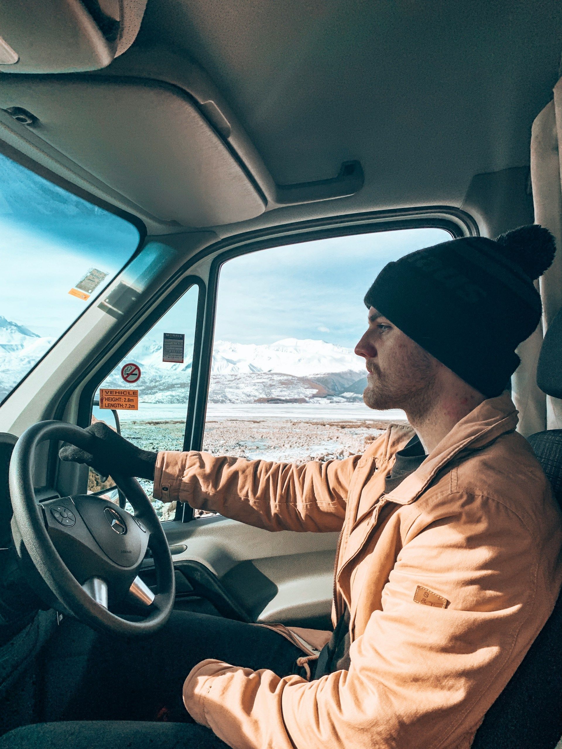 Driver in tan jacket and black beanie driving a van beside snowy mountains.