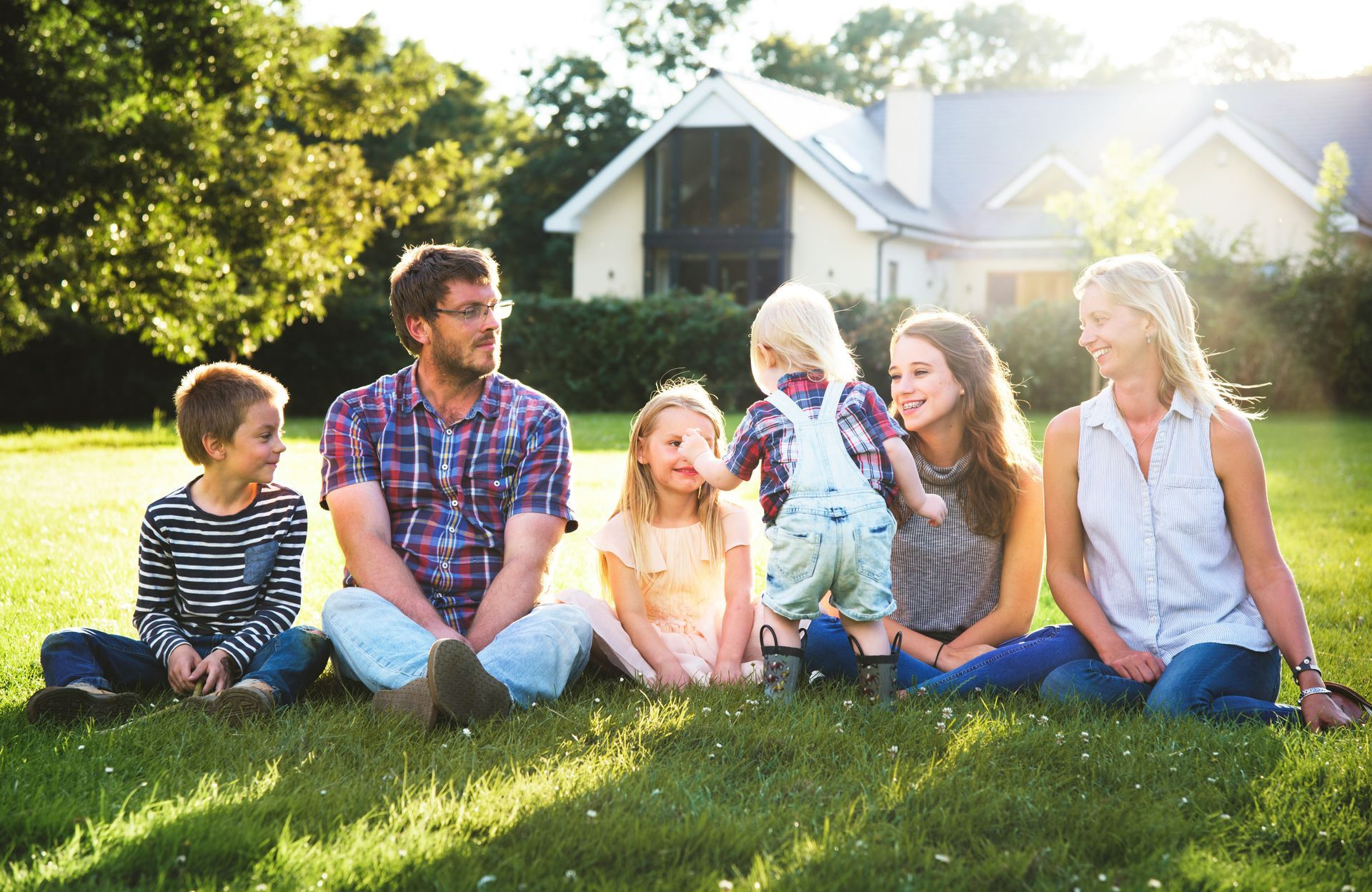 Group of people sitting on grass in a sunny backyard, smiling and chatting together.