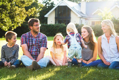 Family sitting on grass in a sunny backyard, smiling and chatting together.