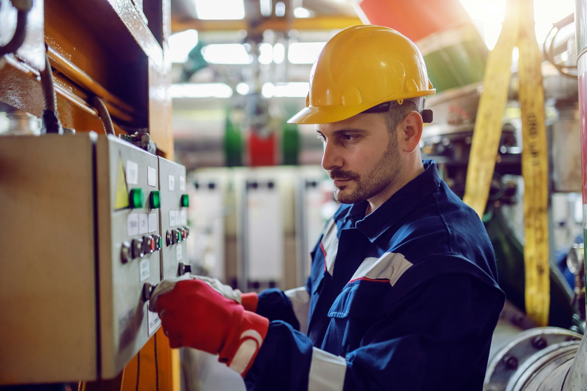 Worker in a yellow hard hat adjusting controls on an industrial machine in a factory.