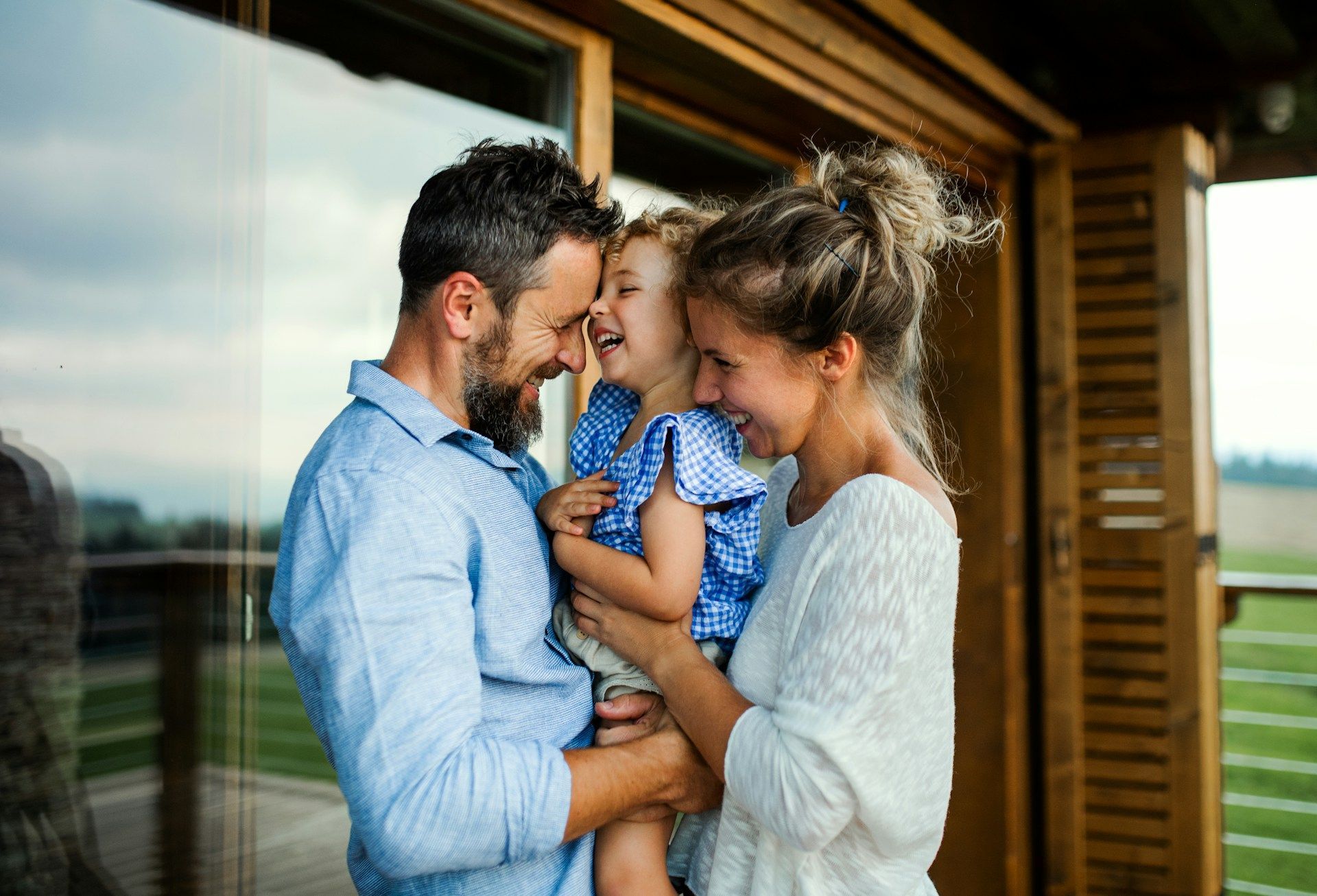 Family smiling on a porch, holding a child in a blue dress beside a wooden house.