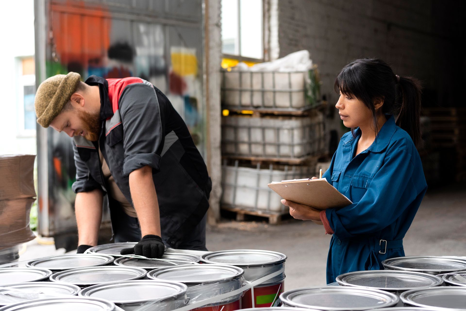 Workers sorting cans in a warehouse, with one writing on a clipboard beside stacked pallets.