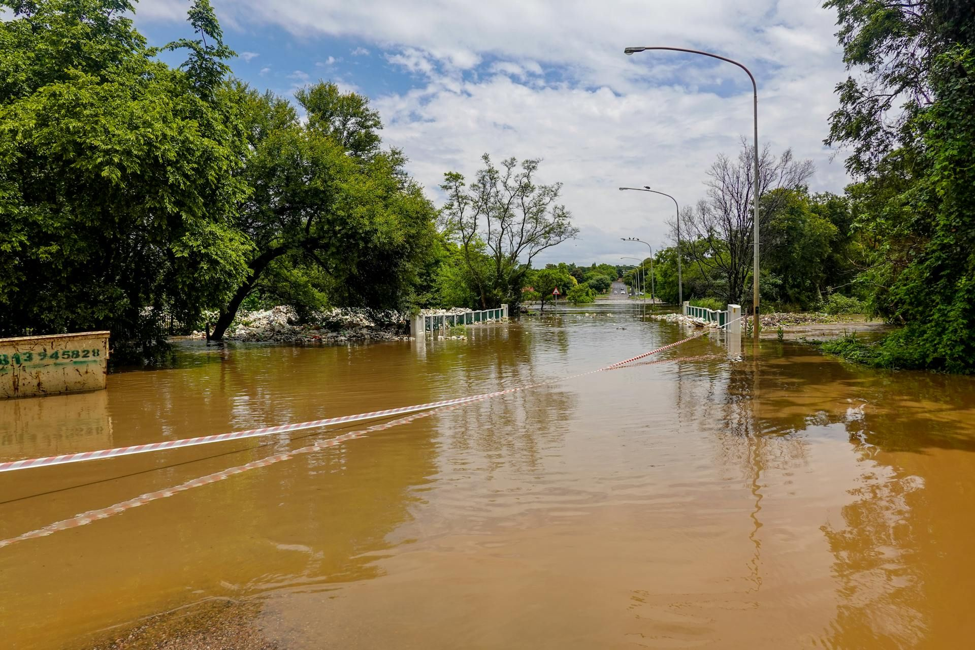 Flooded street with muddy water, trees, and streetlights under a cloudy sky.
