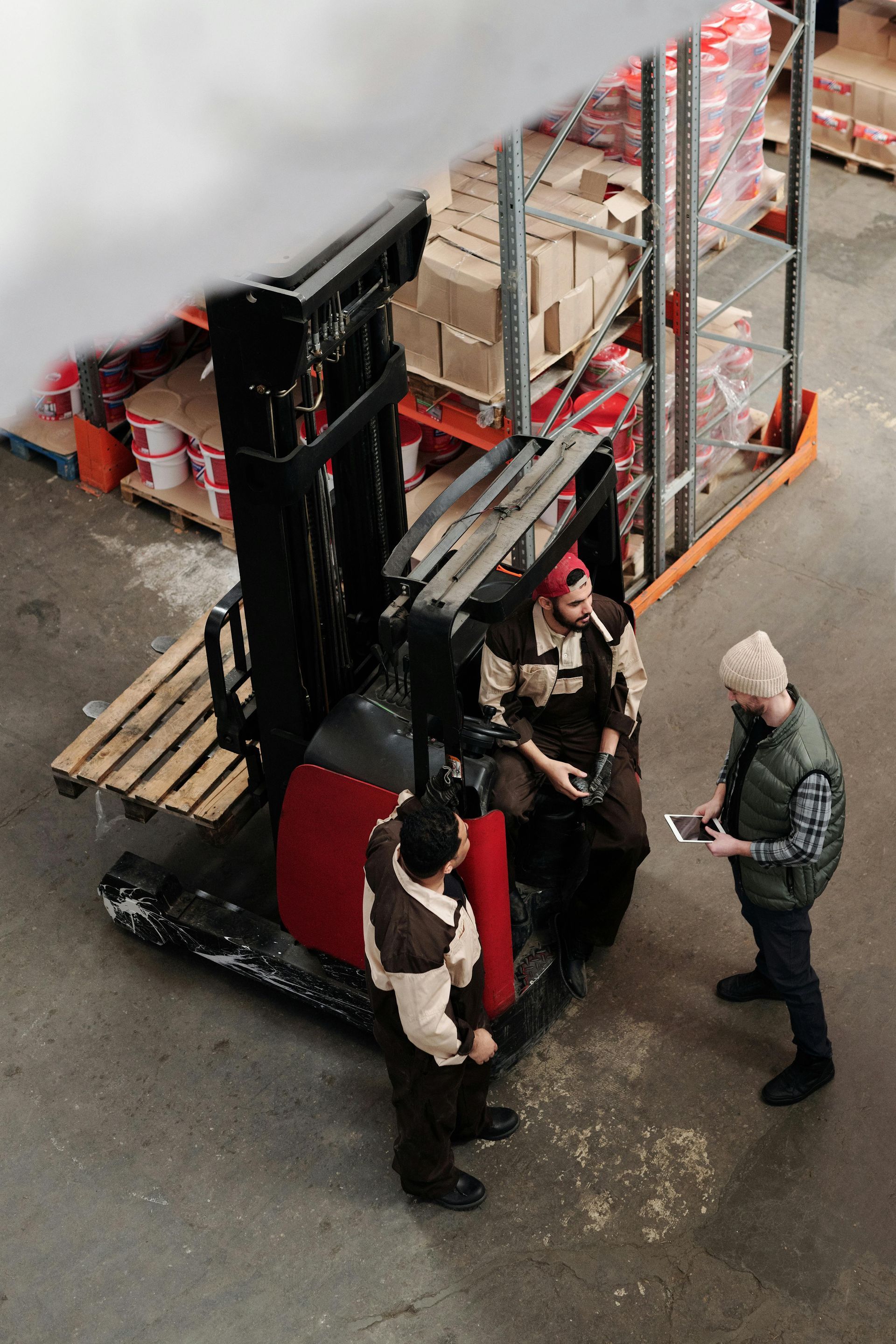 Workers stand beside a red forklift in a warehouse aisle with stacked pallets and shelving.