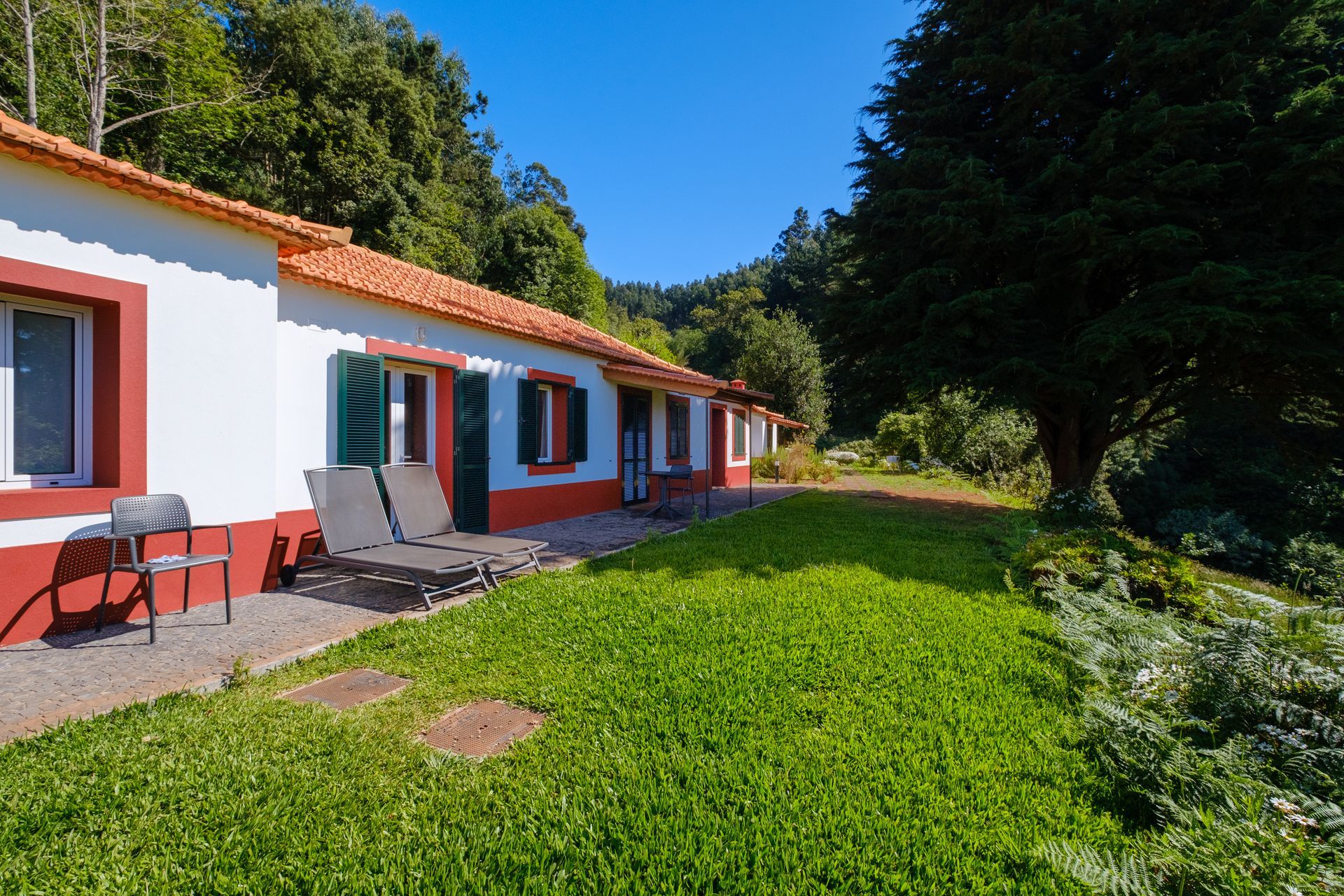 White house with red trim and orange roof, green lawn, two chairs, and trees.