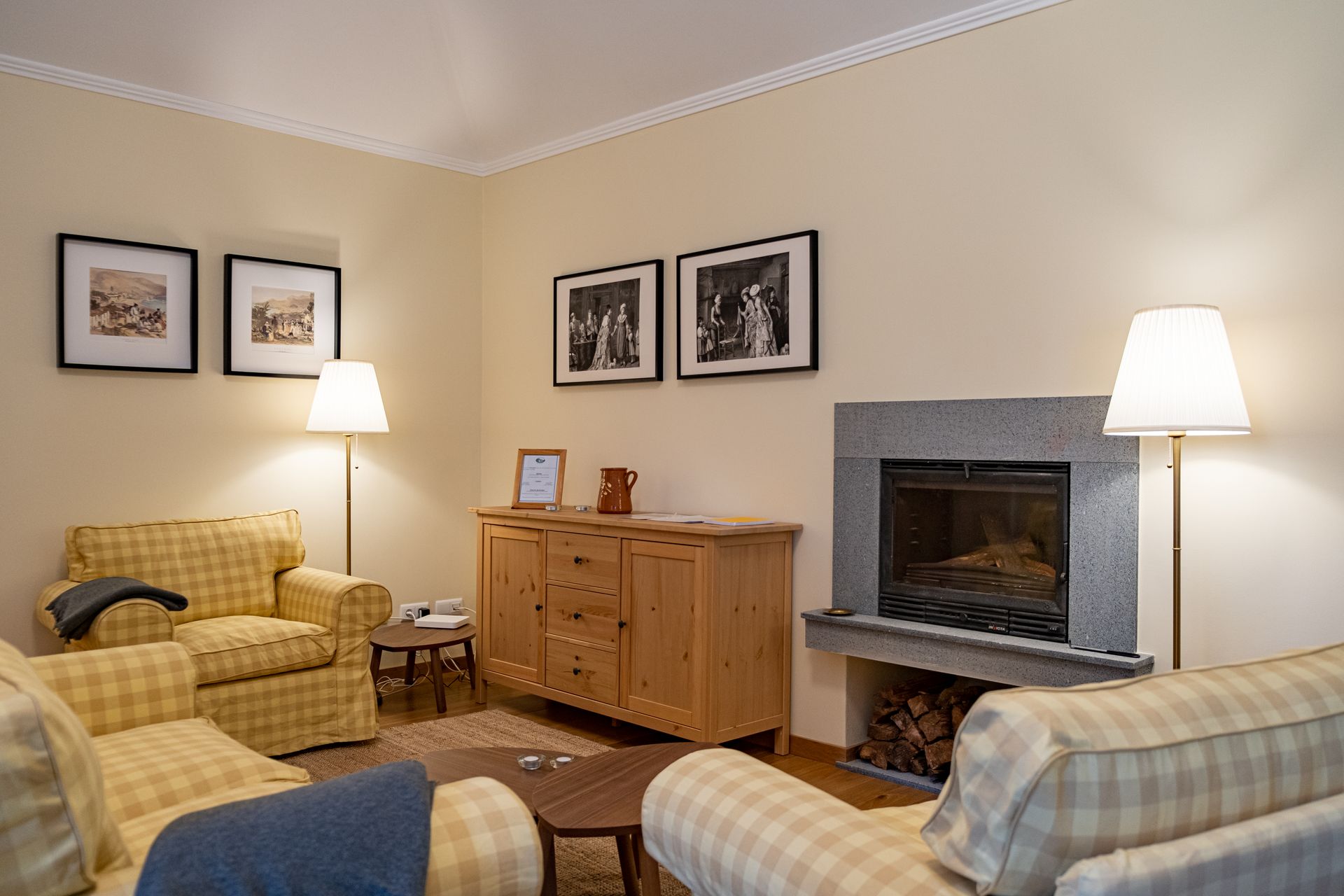 Cozy living room with two plaid sofas, fireplace, wooden cabinet, and framed art. Warm lighting and neutral walls.