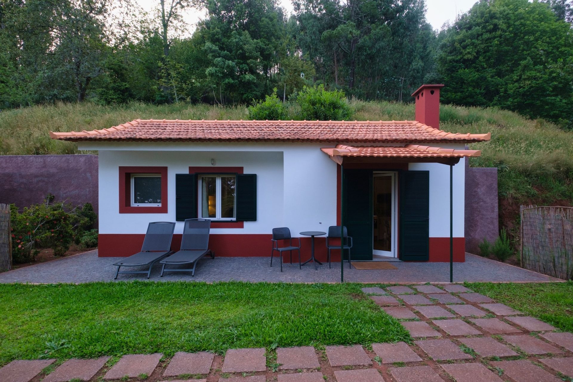 Small white cottage with red trim, a red roof, and black shutters; two lounge chairs are on the patio.