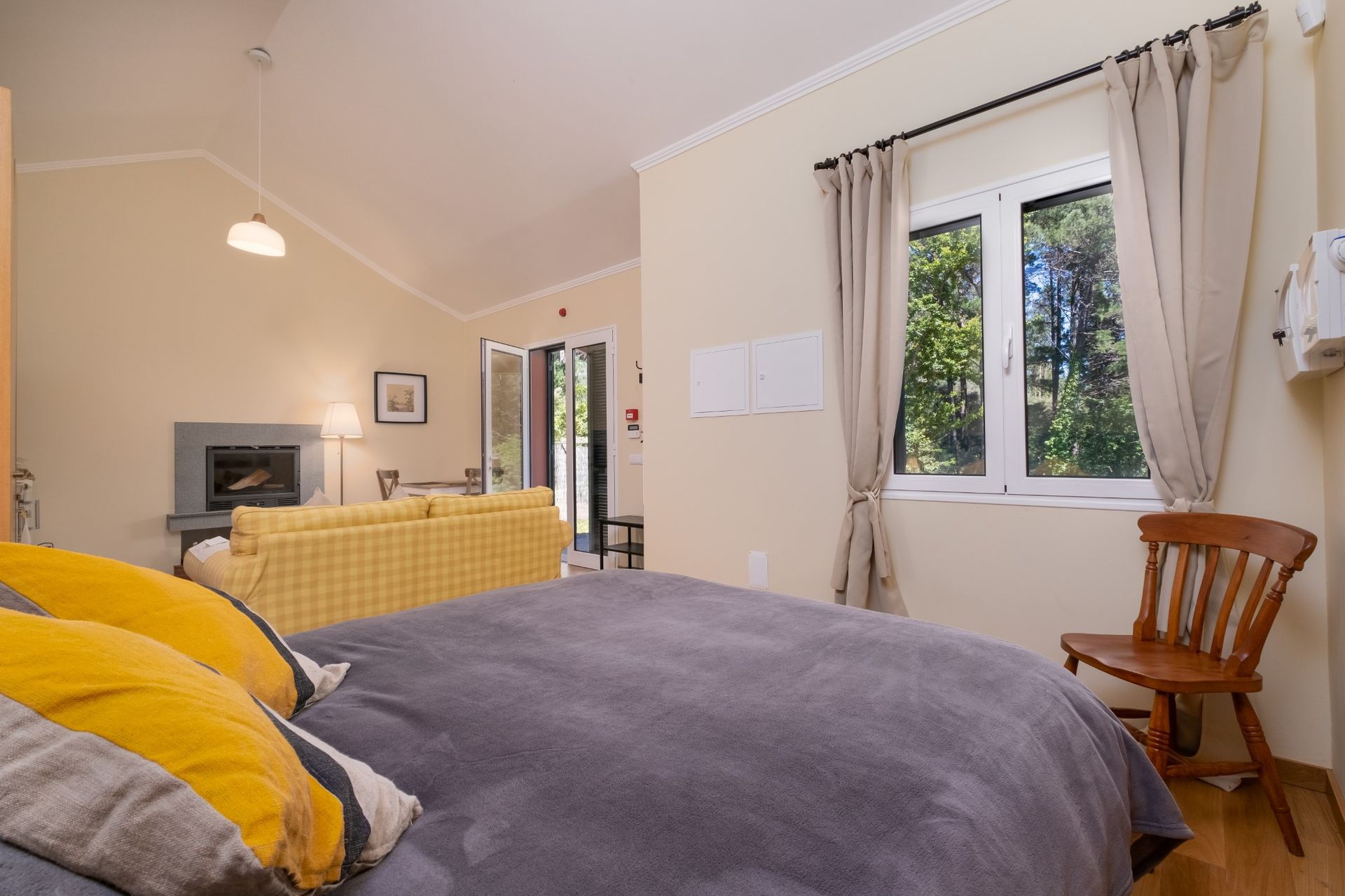Bedroom with gray bed, yellow pillows, wooden chair, window with trees, and sofa.