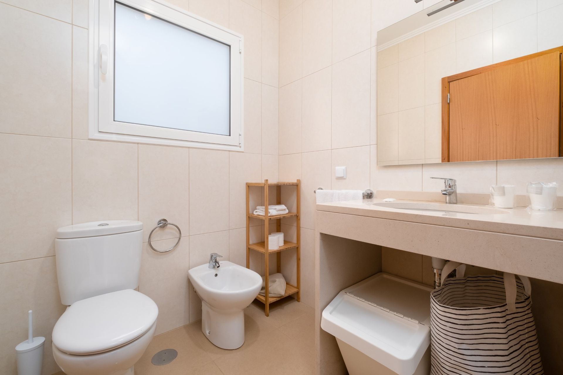 Bathroom with white toilet, bidet, sink, and shelving. Beige walls and floor, window, and mirror.