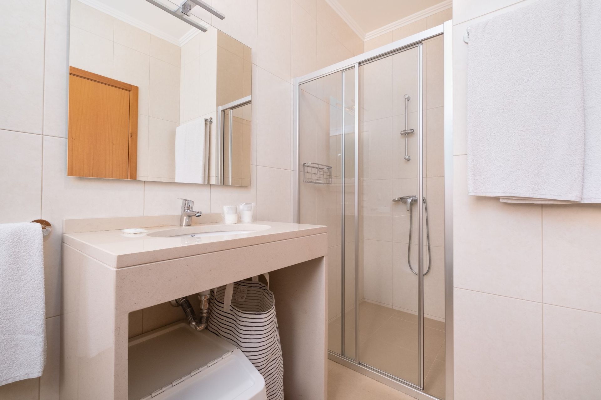 Bathroom with a shower, sink, and beige walls. Towels and a basket are visible.